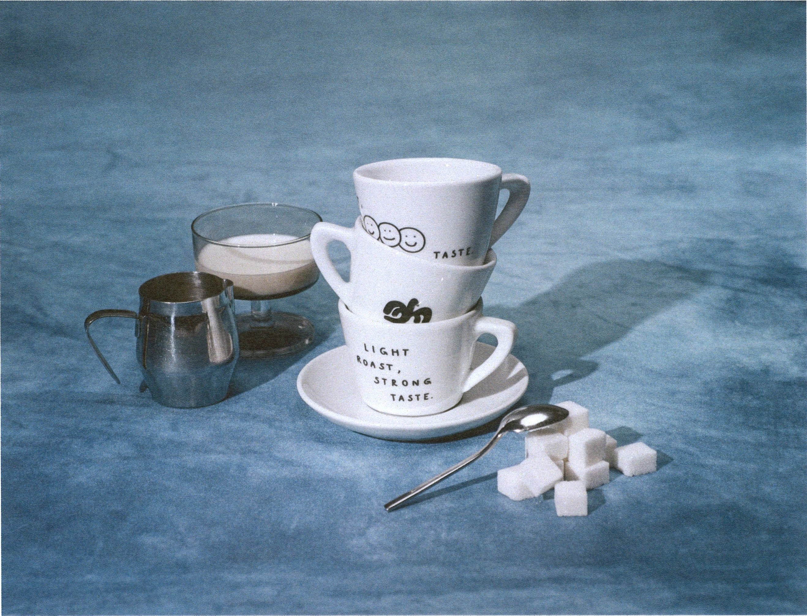 A stack of white ceramic espresso cups and saucers with black printed text and logos, styled on a blue textured surface with a glass of milk, a metal pitcher, a spoon, and sugar cubes.